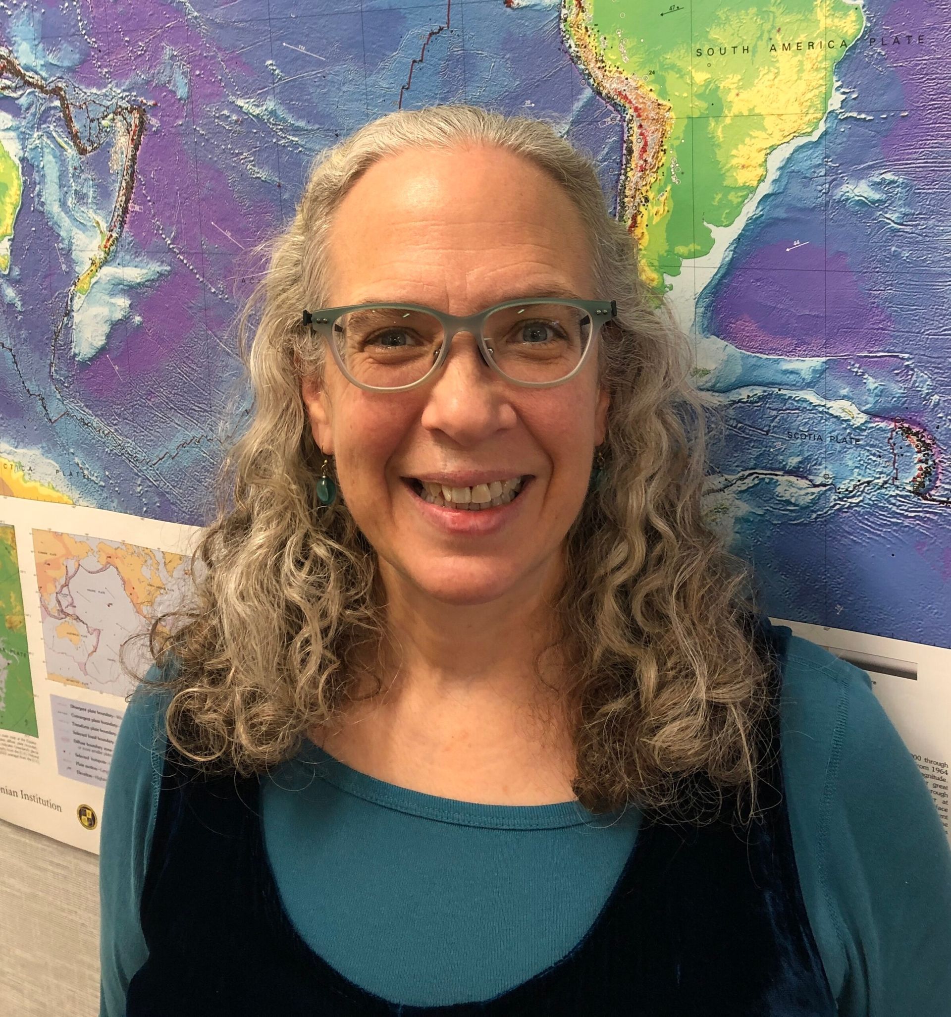 Smiling woman with curly gray hair and glasses, wearing a teal top, standing in front of a colorful map background.