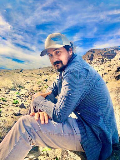 A person in a cap and blue button-down shirt sits in a rocky desert landscape under a blue, cloudy sky.