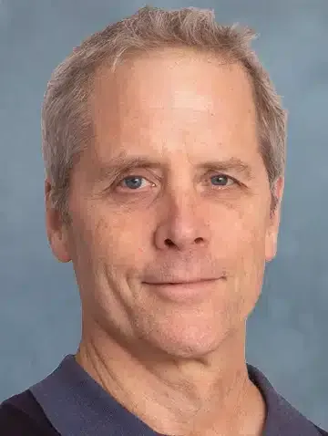 A headshot of a person with short, light hair against a blue-gray background, wearing a dark blue shirt.