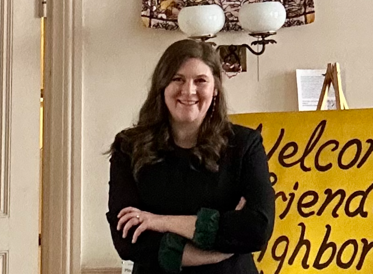 A smiling person with long brown hair stands with arms crossed, wearing a black jacket, in front of a yellow welcome sign.