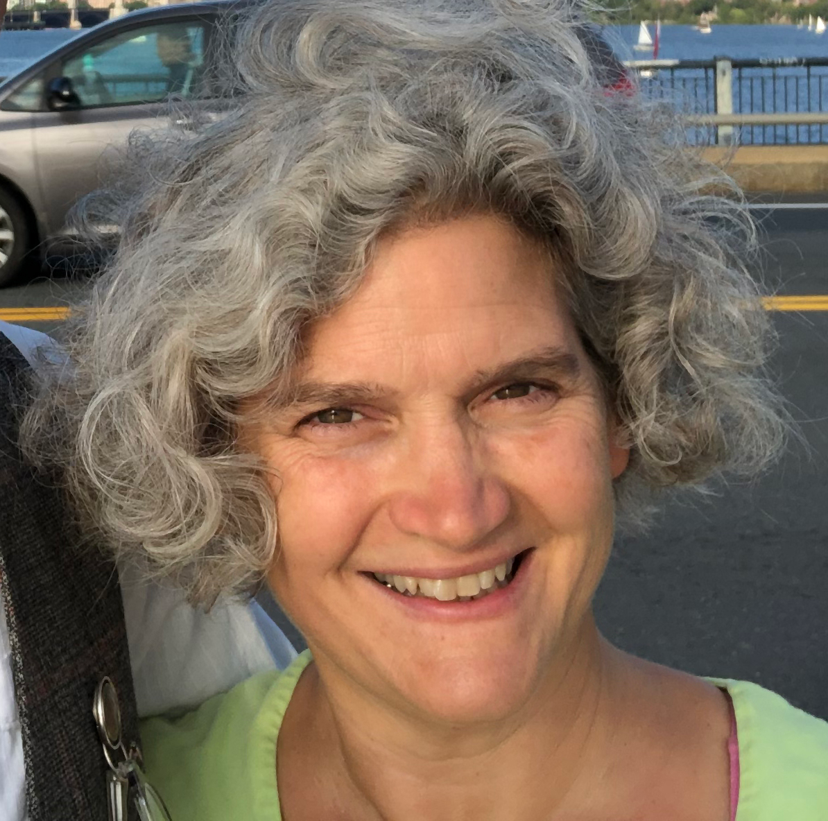 A smiling person with curly gray hair, wearing a light green top, outdoors with a car and water in the background.