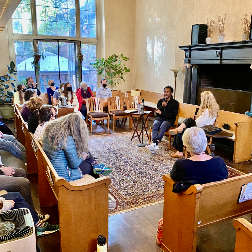 People in room, two speakers at table, seated audience in pews. Window with natural light.