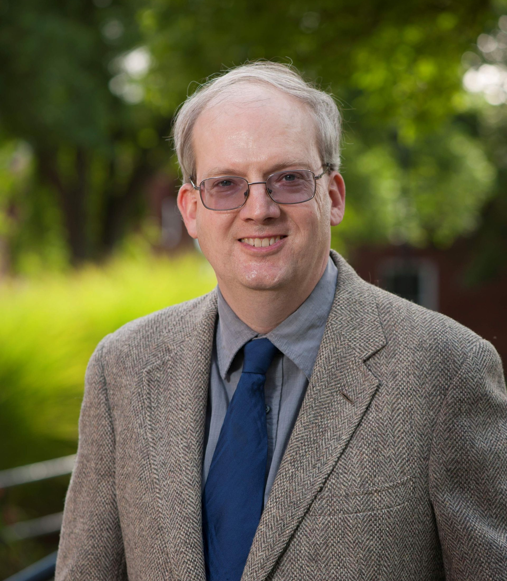 A man wearing a grey shirt, blue tie, and textured tan blazer, standing outdoors with a blurred green background.