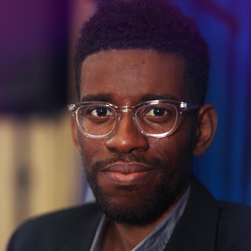 A man with dark hair and a beard wearing clear-framed glasses and a black suit jacket, looking at the camera.