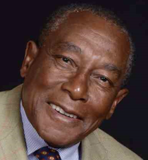 A headshot of a person wearing a tan blazer and a blue striped shirt, smiling gently against a dark background.