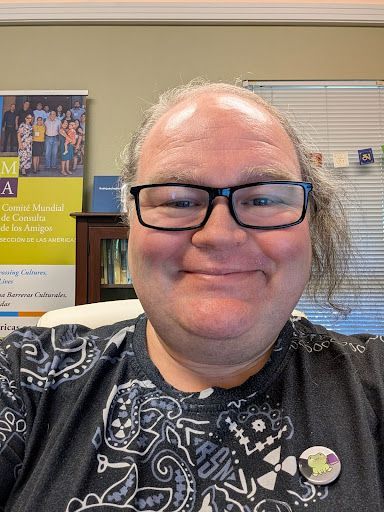 A smiling person with glasses and long, wavy hair wears a black patterned shirt with a pin, posed in an office setting.