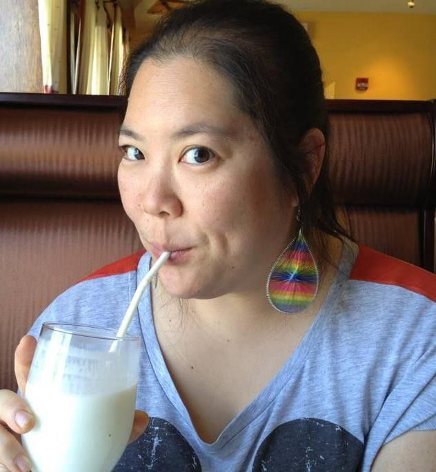 A person with rainbow teardrop-shaped earrings drinks from a glass with a straw while sitting in a restaurant booth.
