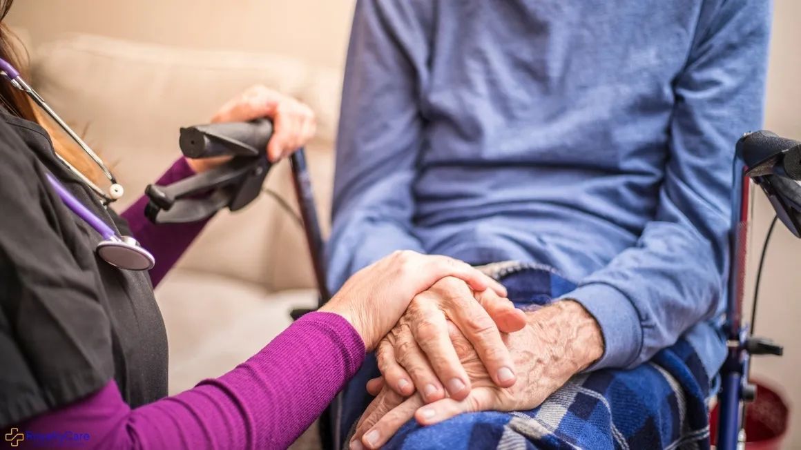Caregiver helping a patient into a wheelchair beside a hospital bed
