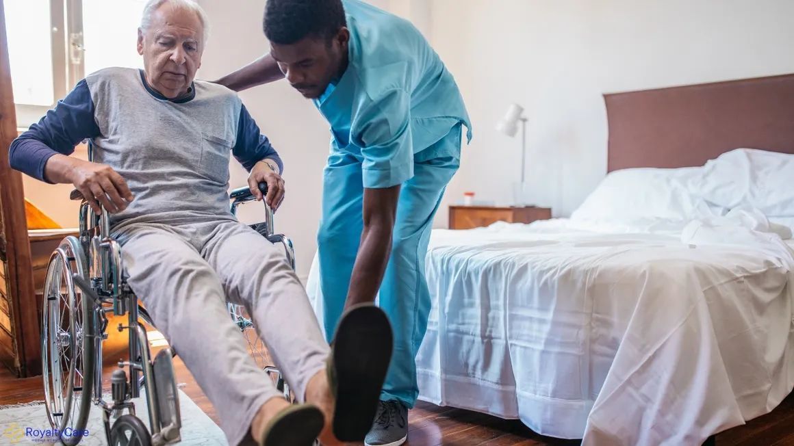 Caregiver helping a patient into a wheelchair beside a hospital bed