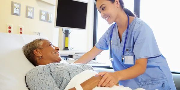 Nurse in blue scrubs smiles while checking on a patient resting in a hospital bed.