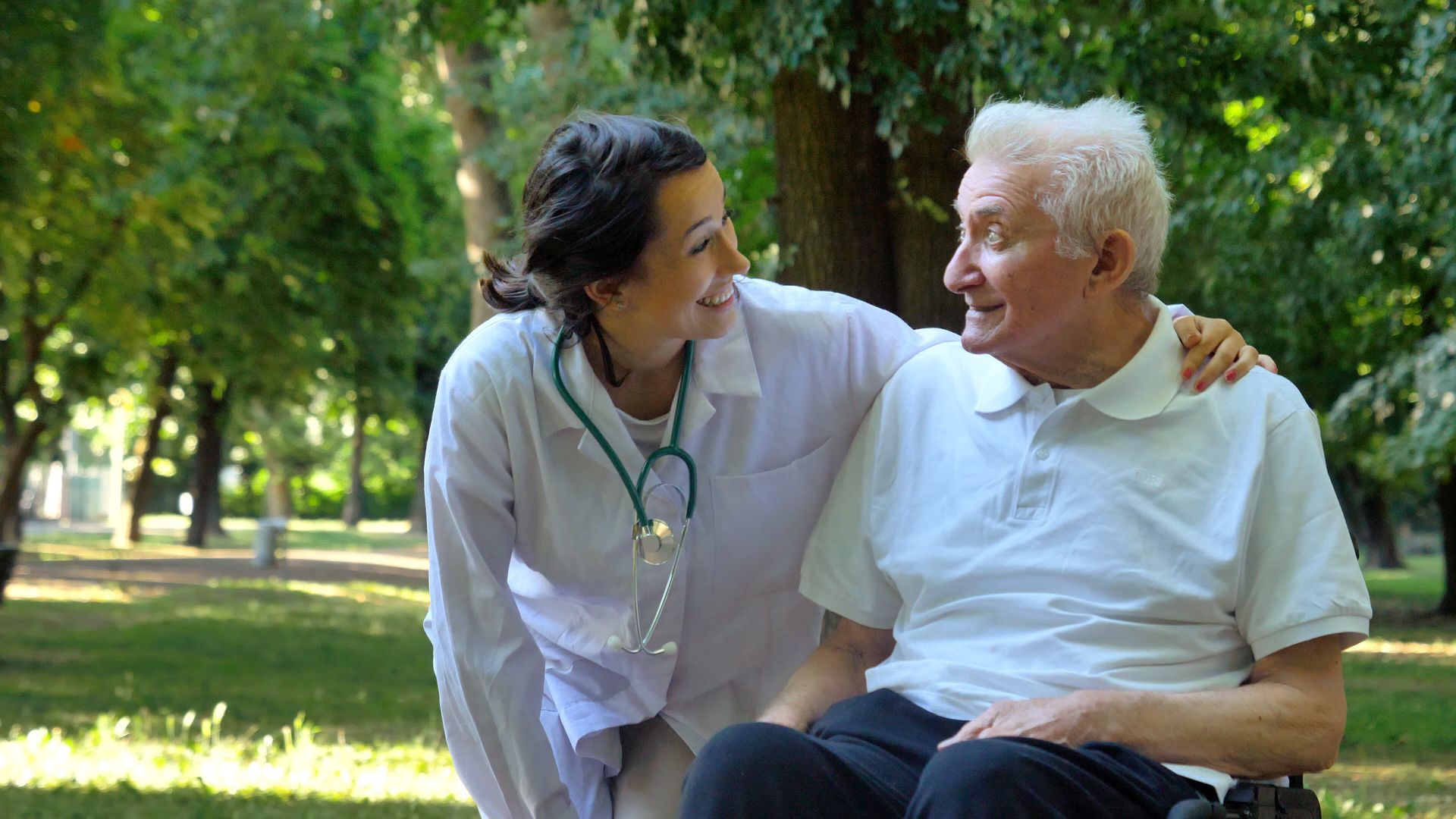 Caregiver leans toward an older man in a wheelchair, smiling in a sunny park.