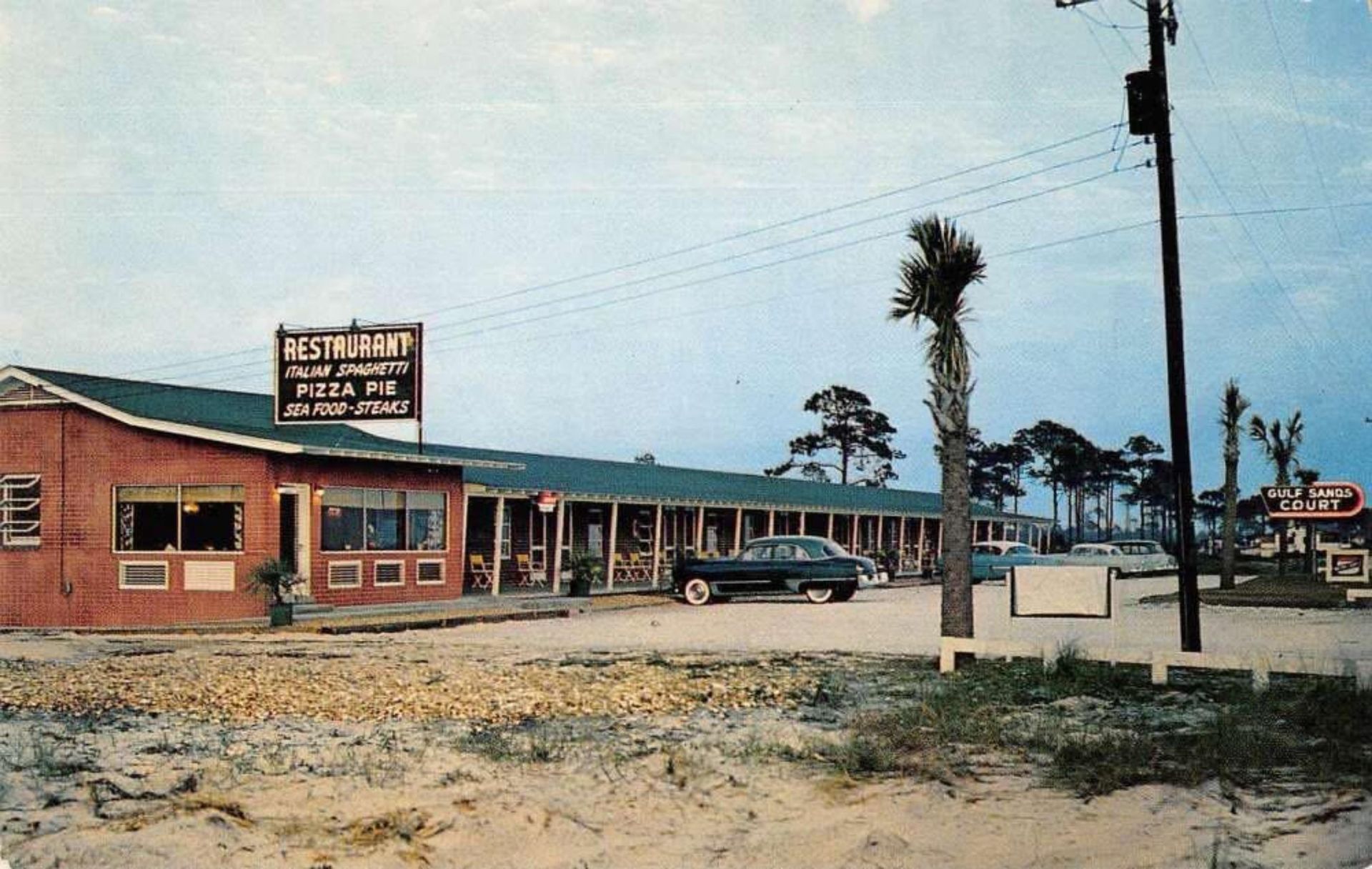 Vintage roadside motel with red brick rooms, blue roof, parked car, and palm trees under a hazy sky