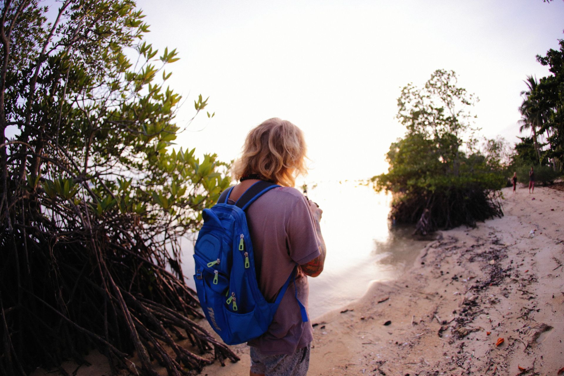 Person with blue backpack standing on a sandy trail by water at sunset