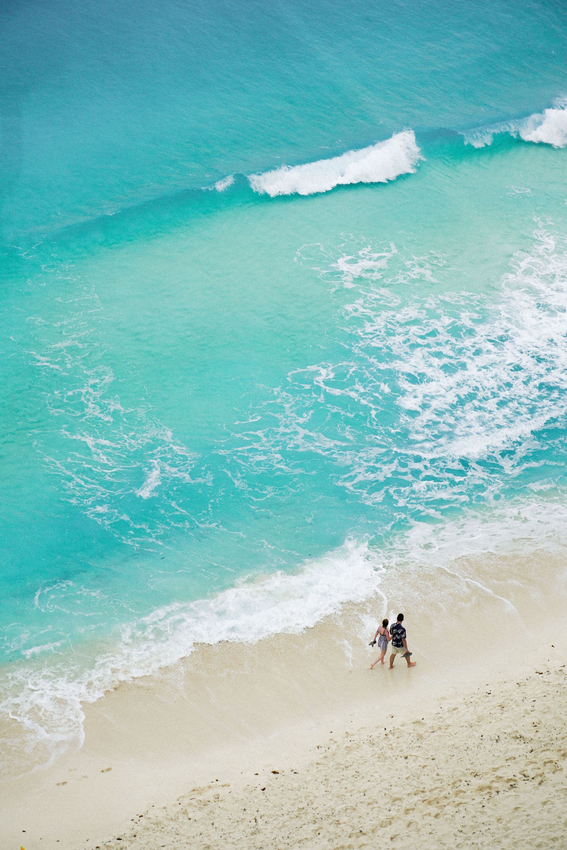 Aerial view of turquoise ocean waves meeting a sandy beach with two people near the shoreline