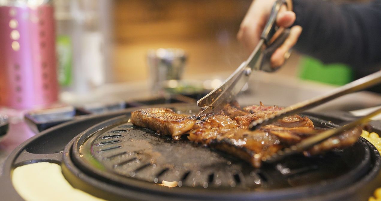 JPOT BBQ person grilling pork on a table grill using kitchen scissors and metal tongs