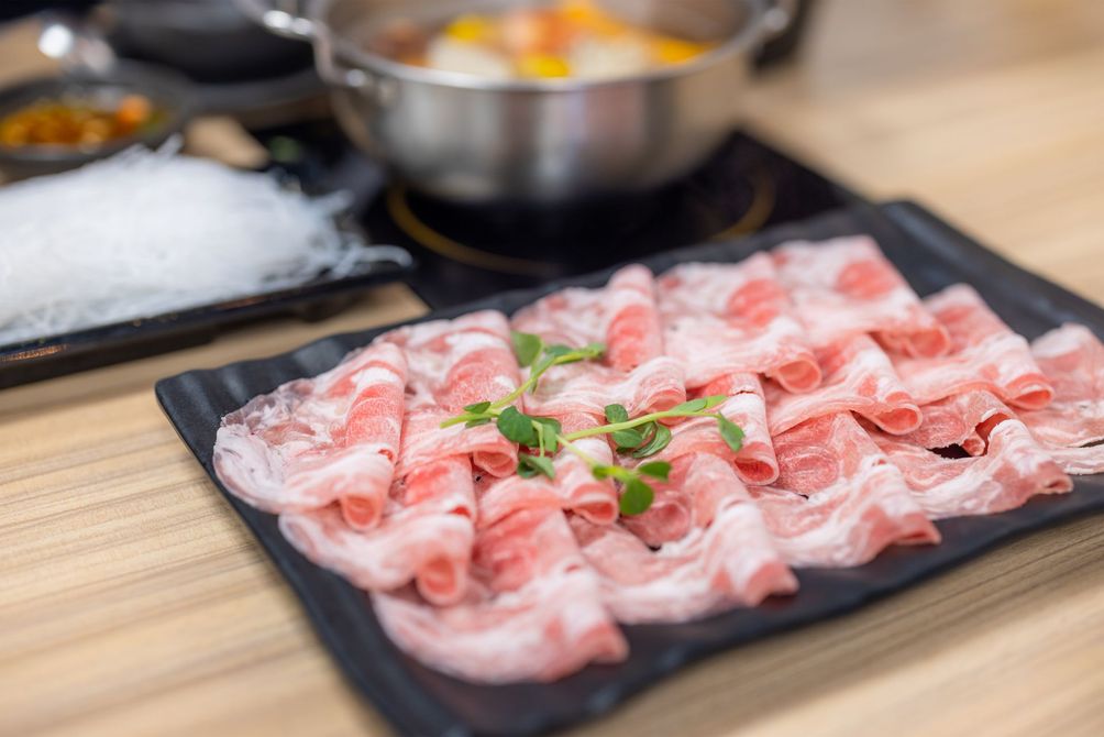 JPOT Japanese Hot Pot raw sliced pork meat on a black plate with other hot pot food items and soup broth pot in background