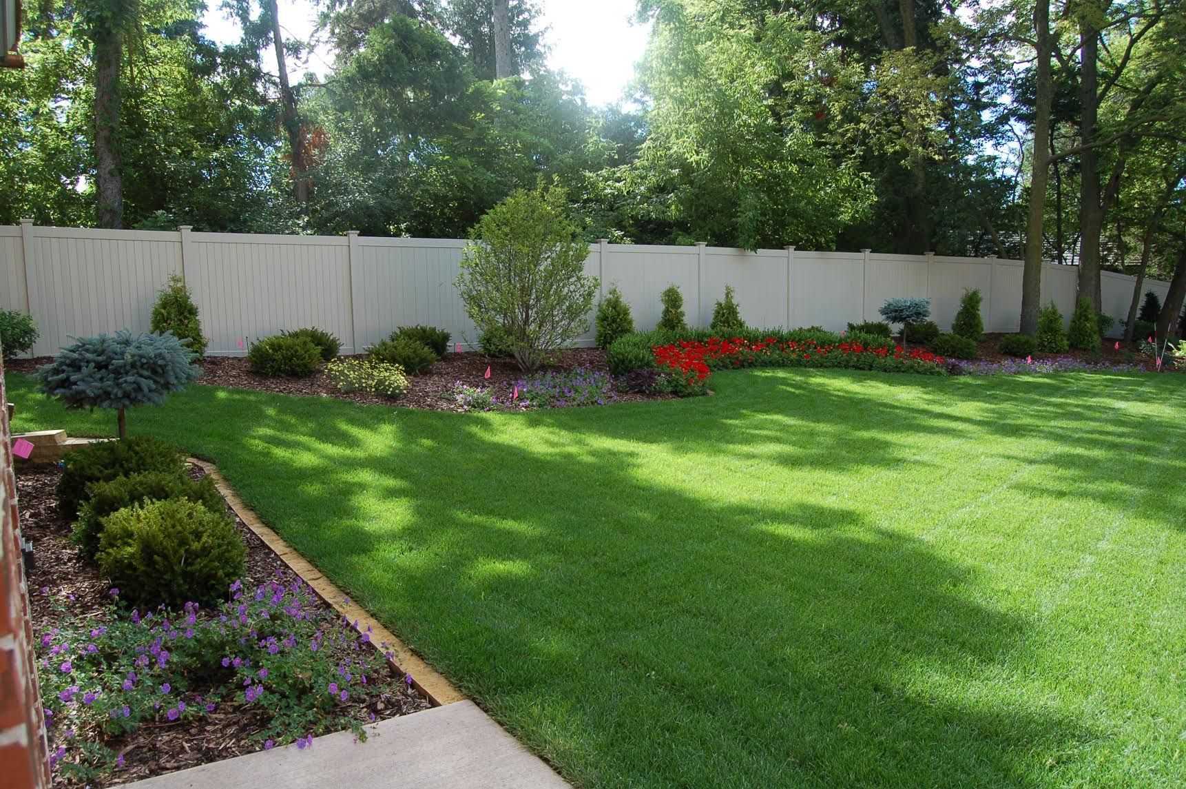A manicured green lawn framed by a curved garden bed with shrubs, purple flowers, and a red flower border by a white fence.
