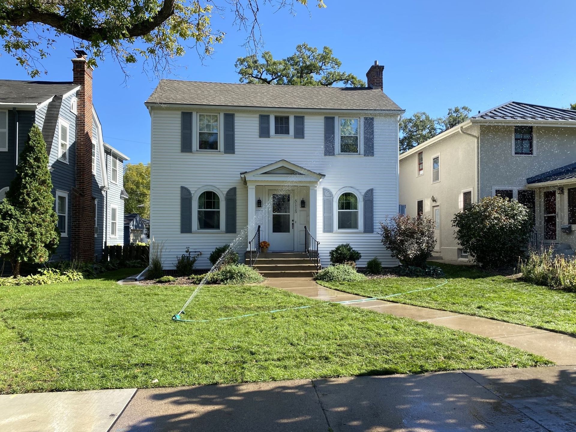 A two-story white house with blue shutters stands between two neighboring homes under a clear blue sky.