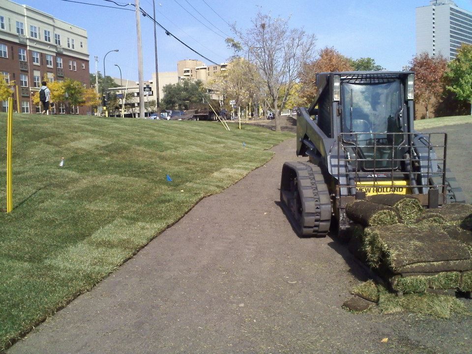 A skid-steer loader sits on a paved path next to a newly sodded grassy slope on a sunny day.