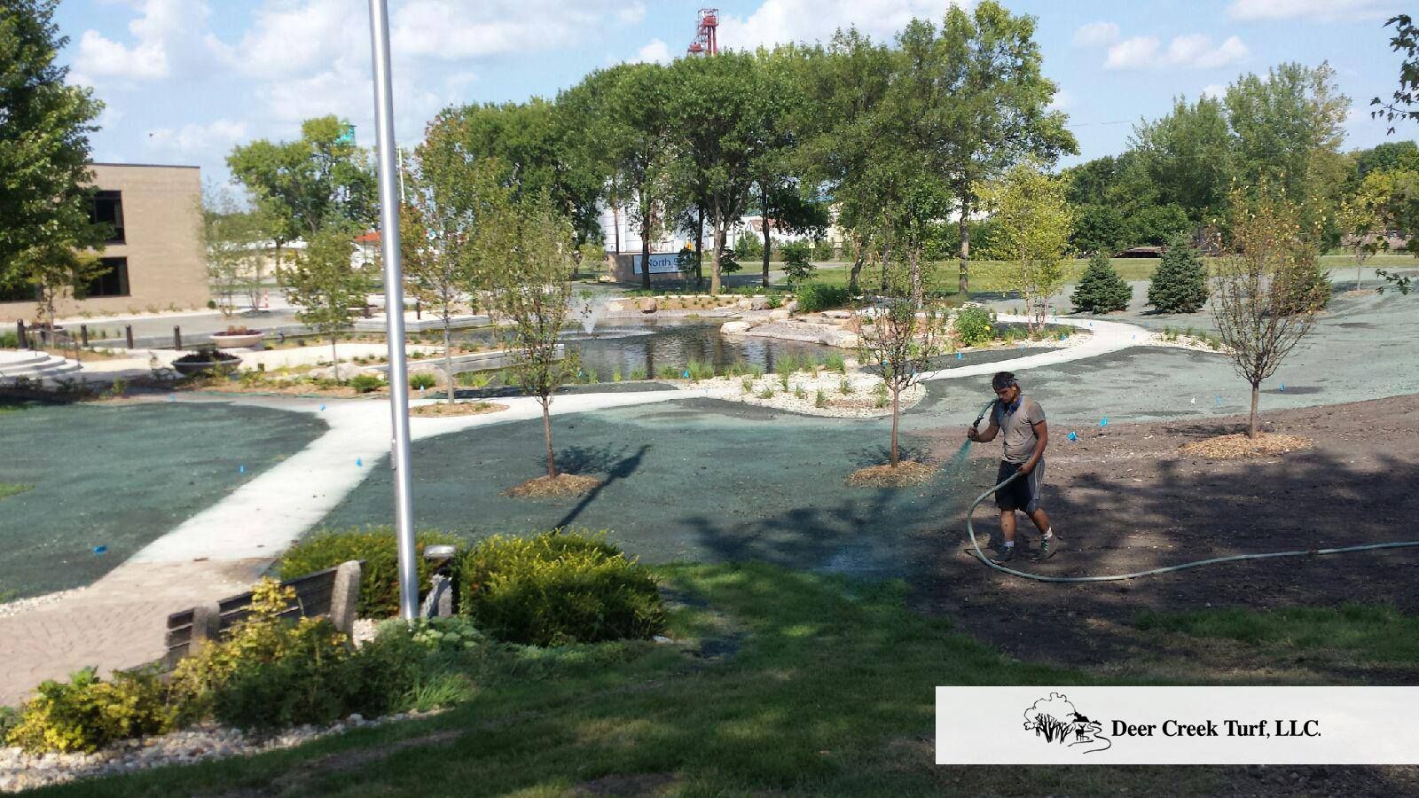 A person sprays green mulch over a newly landscaped park area with trees, a small pond, and a stone walkway.