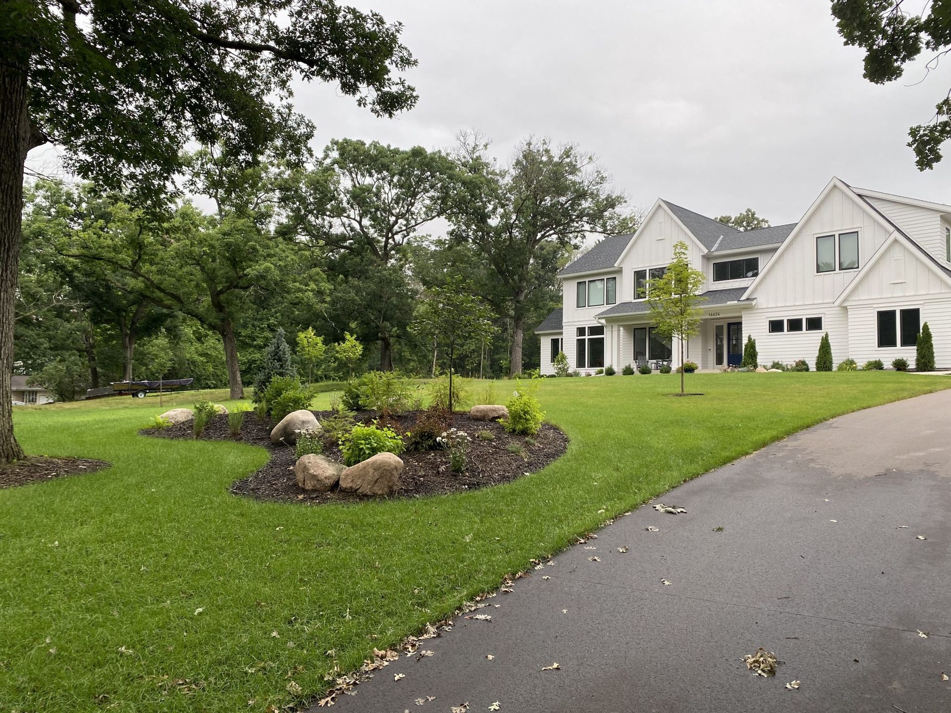 A large, two-story white modern farmhouse with a dark roof sits behind a lush green lawn, trees, and a paved driveway.