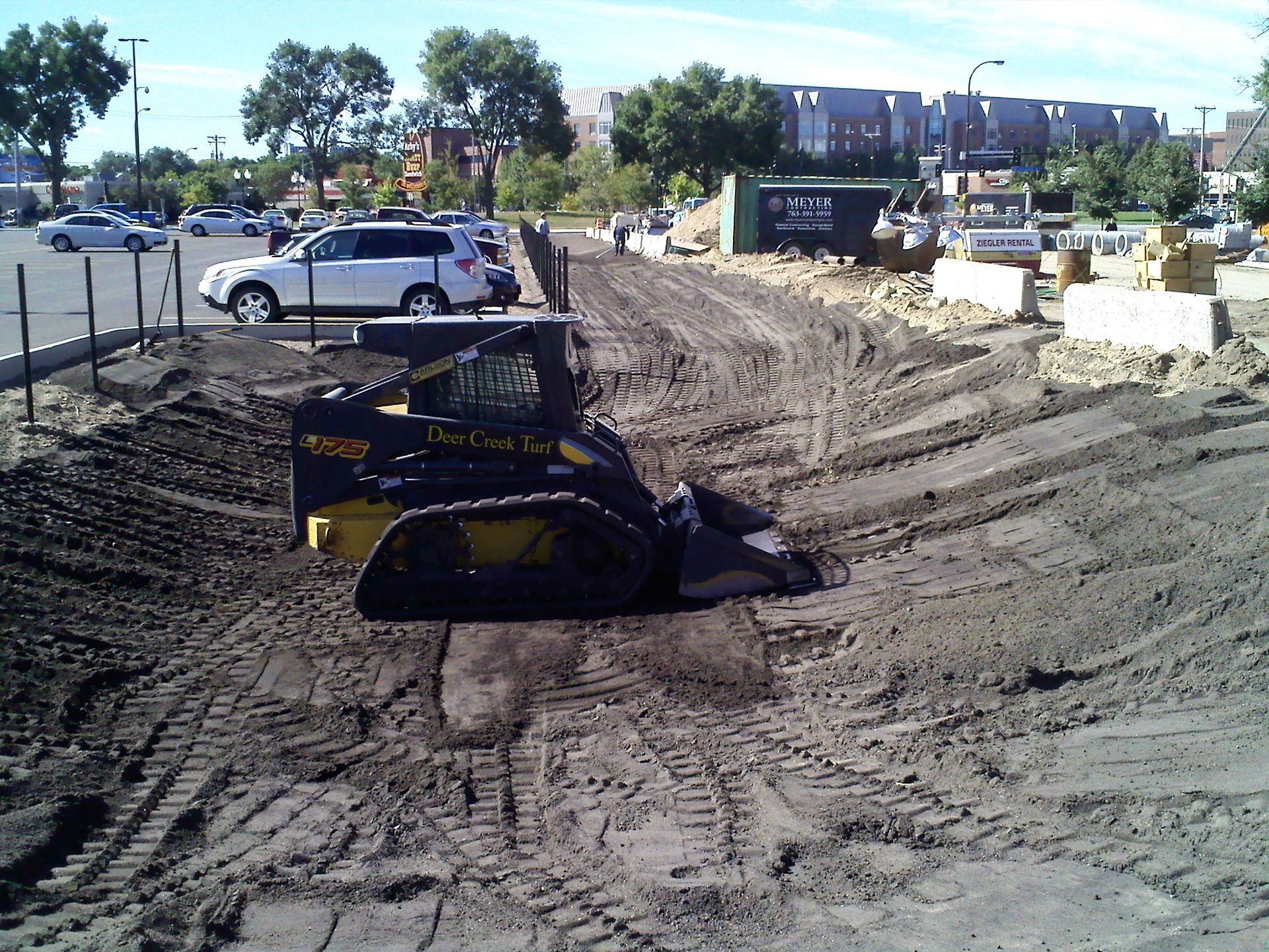 A yellow and black skid-steer loader sits on a dirt construction site near a parking lot.