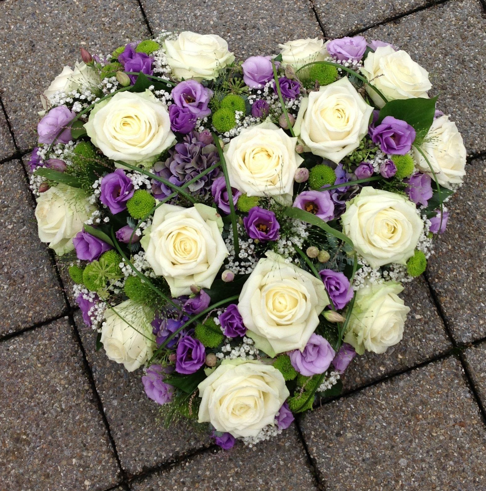 Ein herzförmiges Arrangement aus weißen und violetten Rosen auf einem gepflasterten Gehweg.
