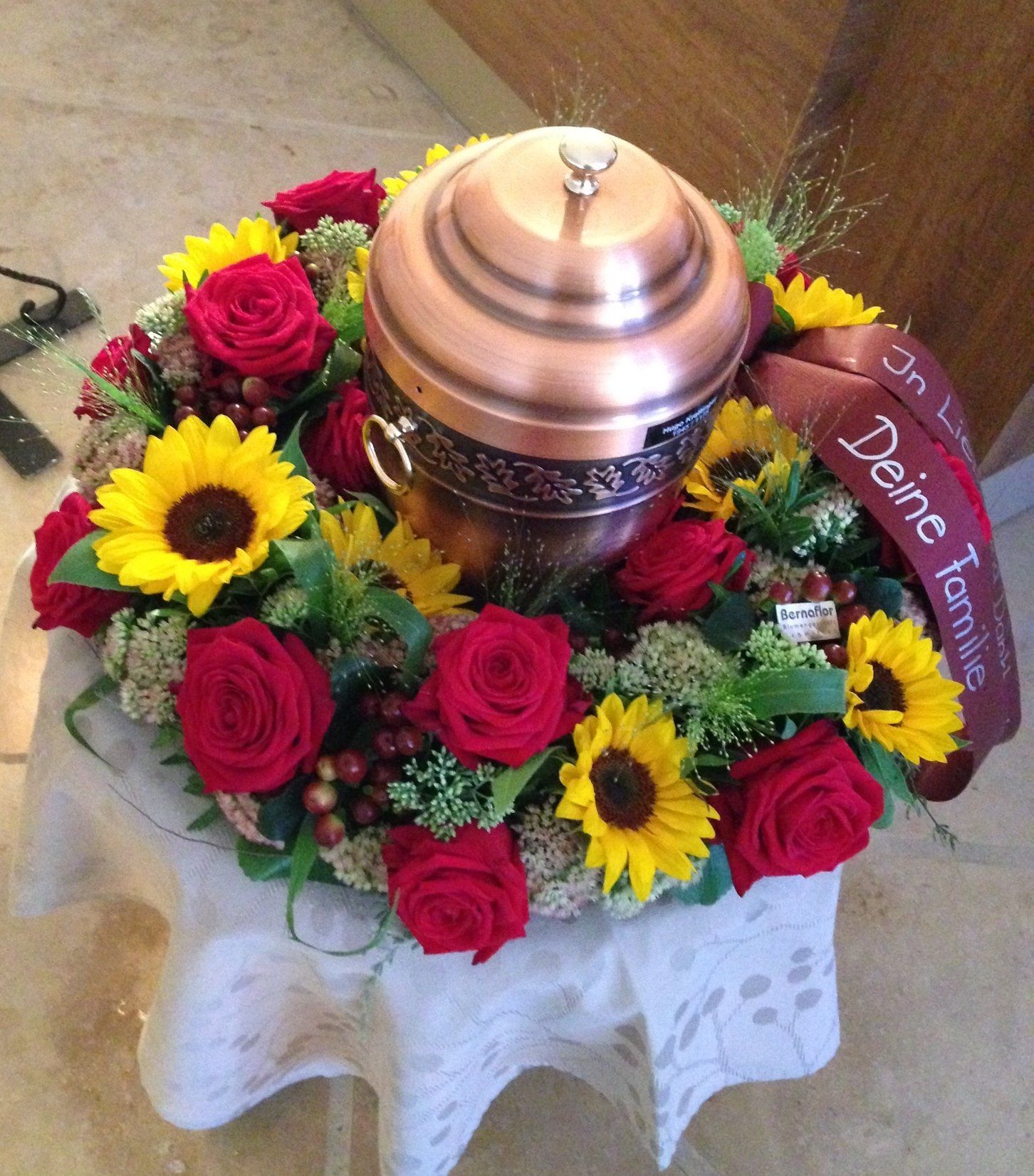 Eine Kupferurne, umgeben von roten Rosen und gelben Sonnenblumen