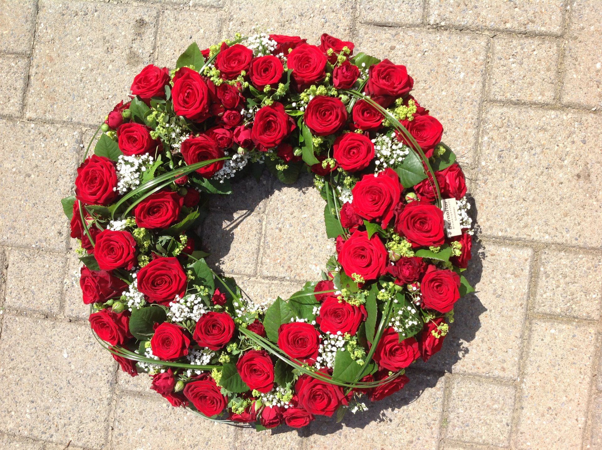 Auf einem gepflasterten Gehweg liegt ein Kranz aus roten Rosen.