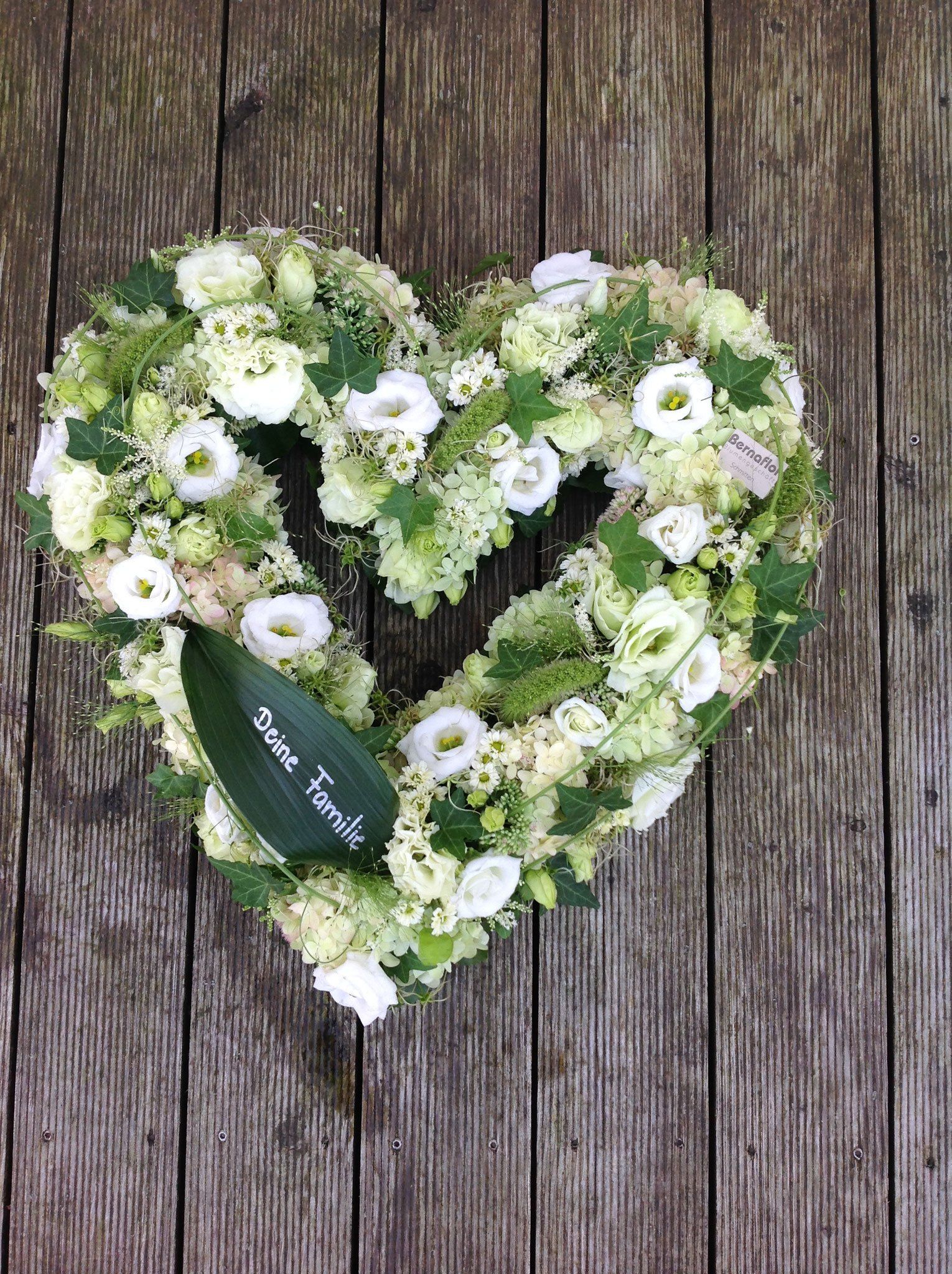 Auf einem Holztisch steht ein herzförmiger Kranz aus grünen und weißen Blumen.