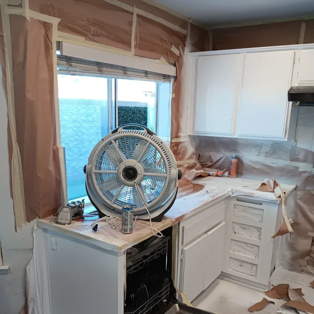 Kitchen under renovation with white cabinets, exposed walls, and a fan by the window overlooking the ocean