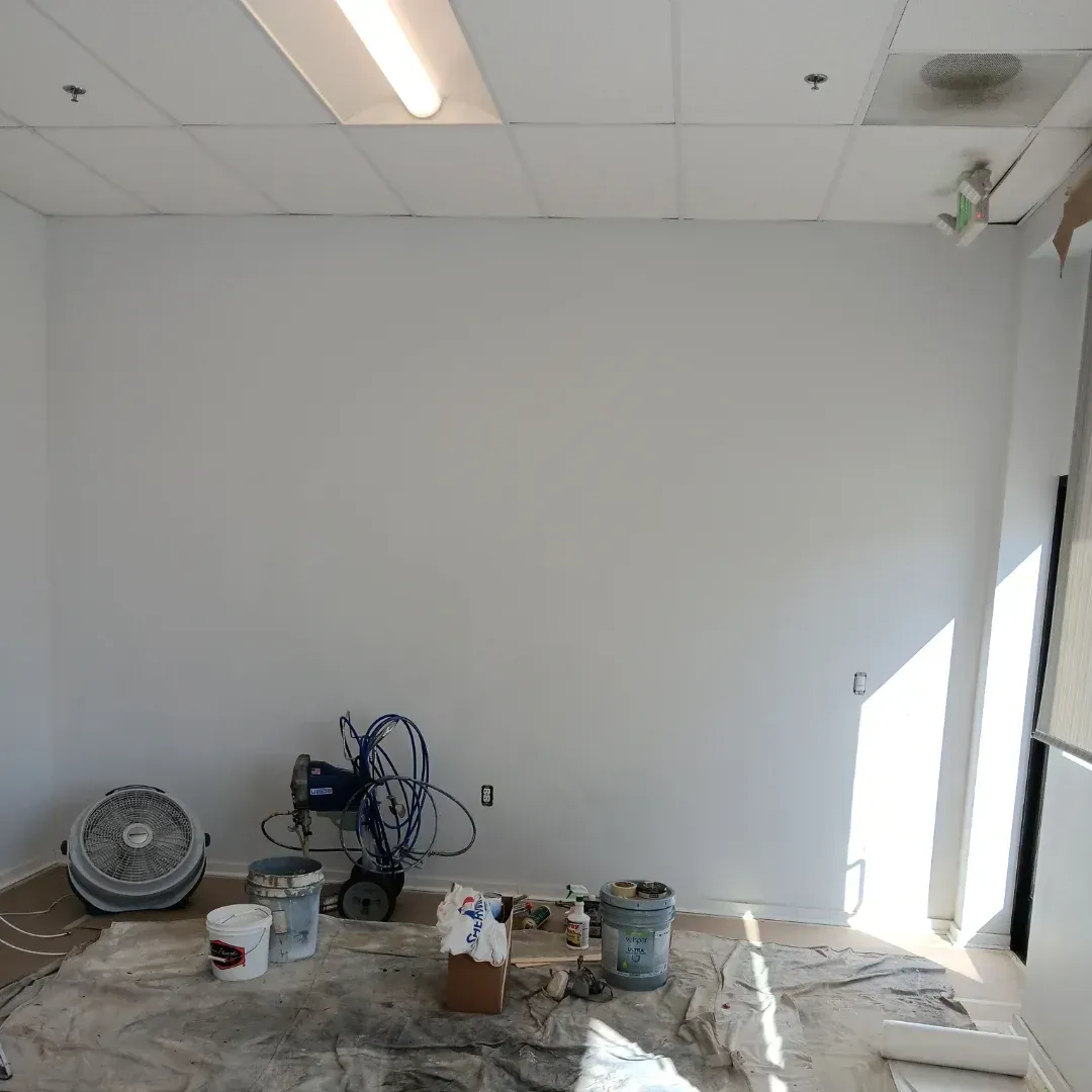 Empty room under renovation with paint supplies, tools, and protective covering on the floor.