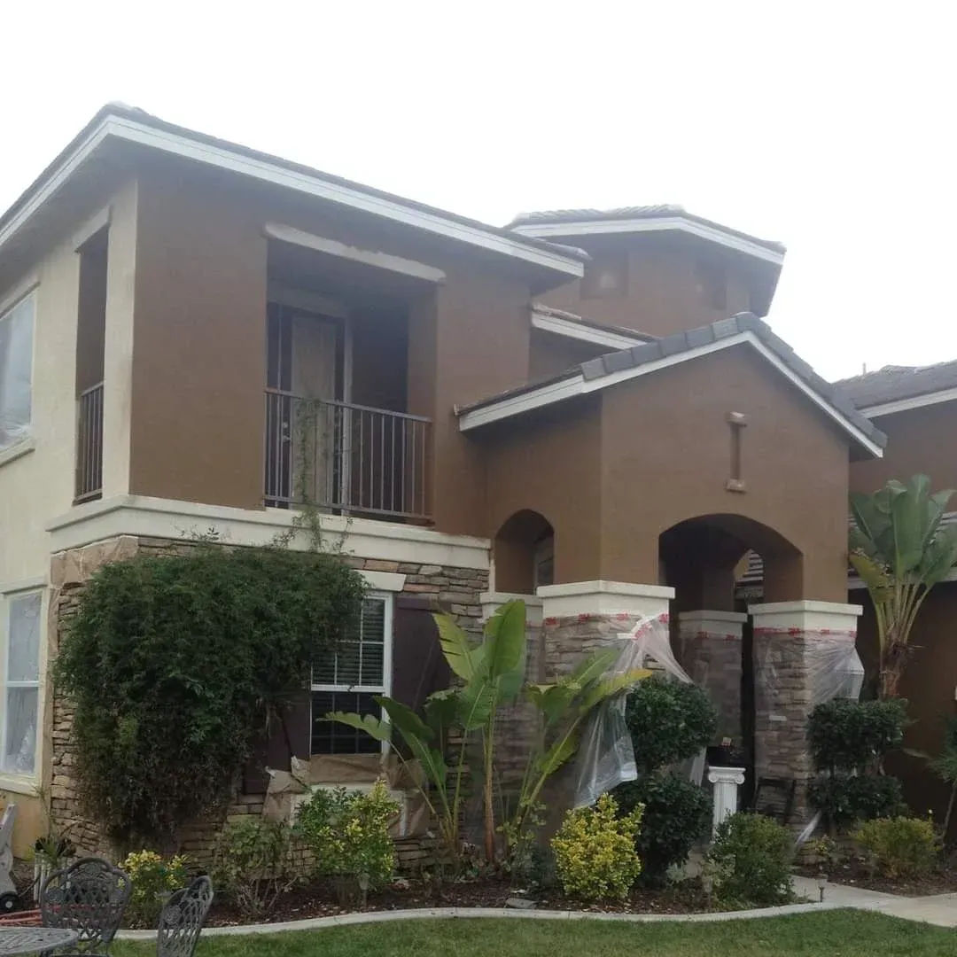 Two-story tan and brown house with a balcony, arched entryway, and lush landscaping