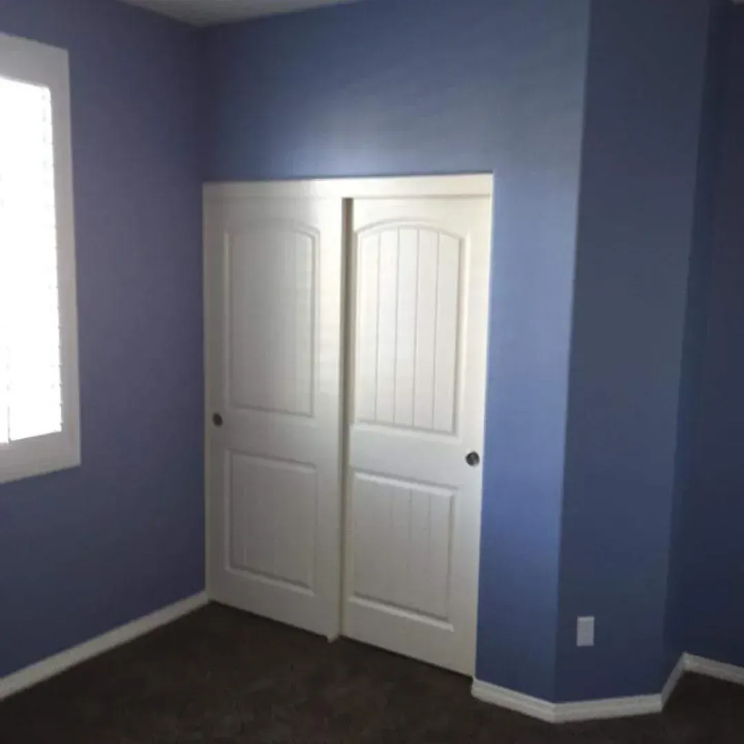 Blue bedroom with white double closet doors and dark carpet, lit by a window on the left.