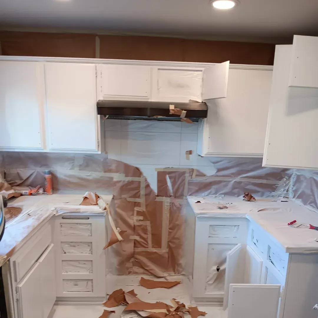 Kitchen under renovation with white cabinets covered in plastic and masking tape, debris on the floor.