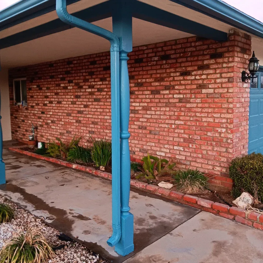 Covered walkway with blue columns beside a red brick wall and small plants along the edge.