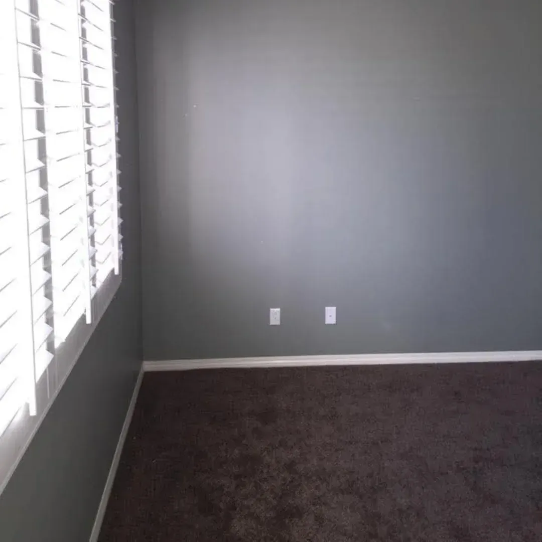 Empty gray room with dark carpet, white trim, and two wall outlets near a window on the left.