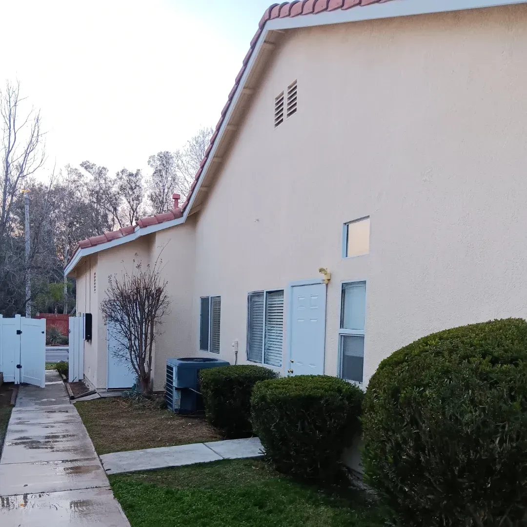 Suburban townhouse exterior with beige stucco walls, white shutters, trimmed shrubs, and a sidewalk