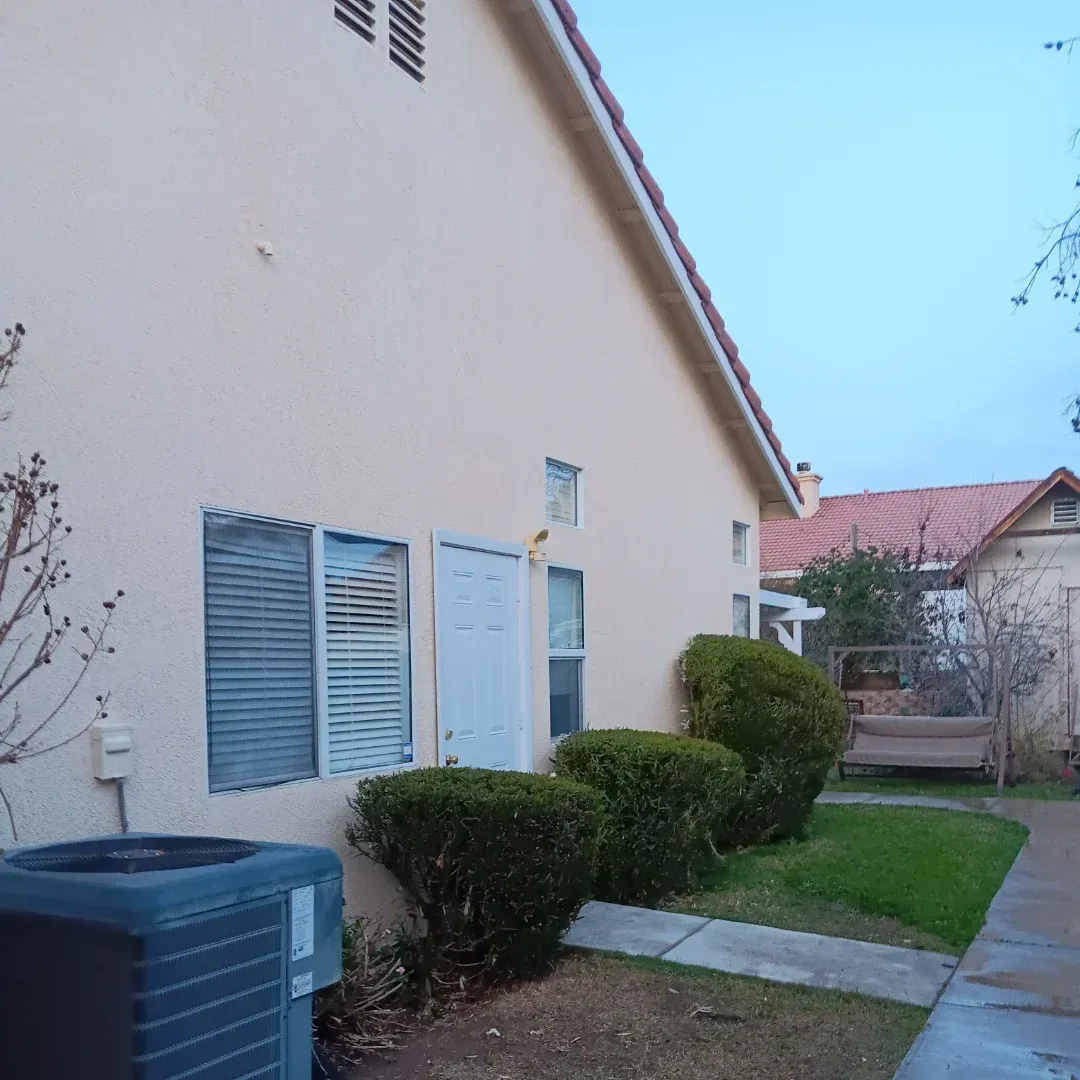 Side view of a beige house with windows, shrubs, a walkway, and an outdoor AC unit in a suburban yard