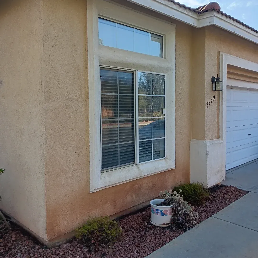 Front exterior of a tan house with a white-framed window, garage door, and small rock landscape bed.