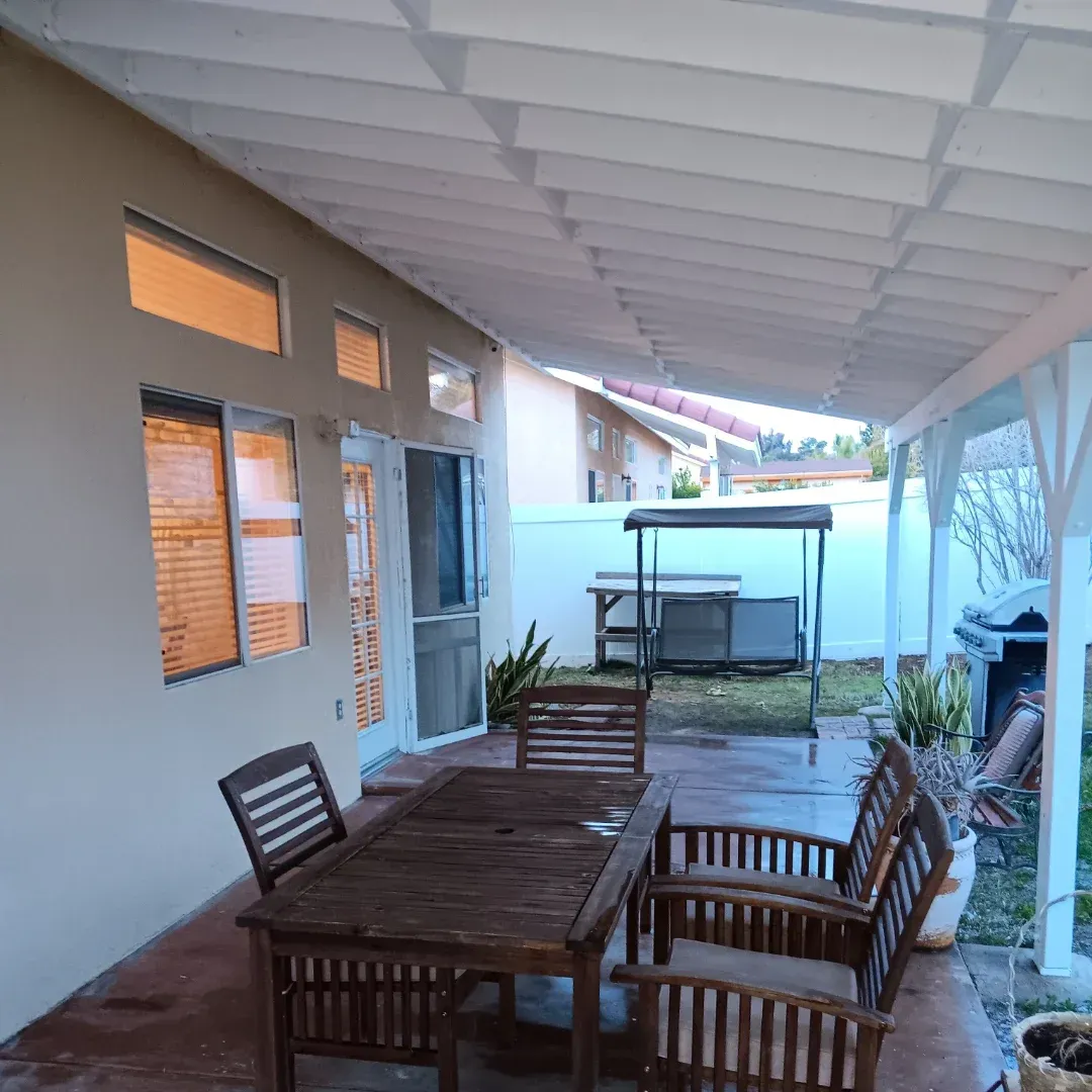 Covered patio with wooden table and chairs beside a white house and garden area