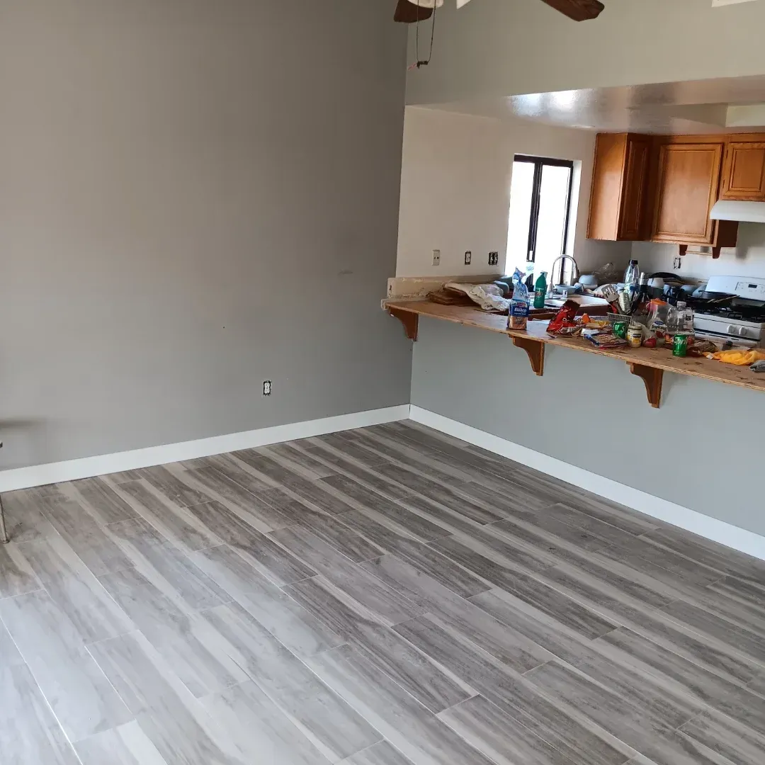 Empty living room with gray wood-look flooring and a kitchen pass-through cluttered with items.