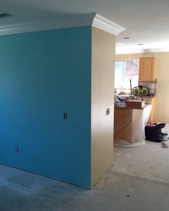 Partially painted room with bright blue wall, beige corner, and a view into a kitchen area.