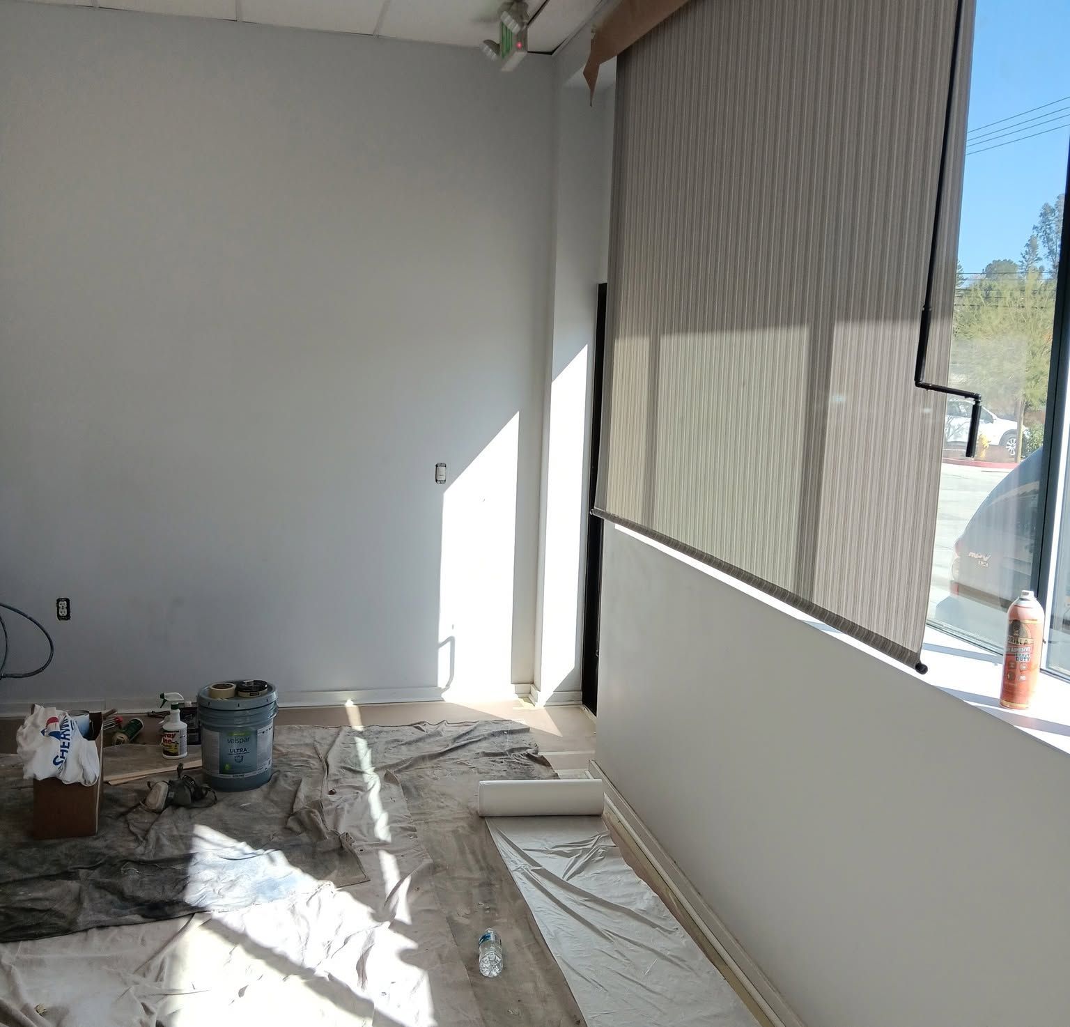 Interior room under renovation with covered floor, white walls, paint buckets, and a large shaded window.