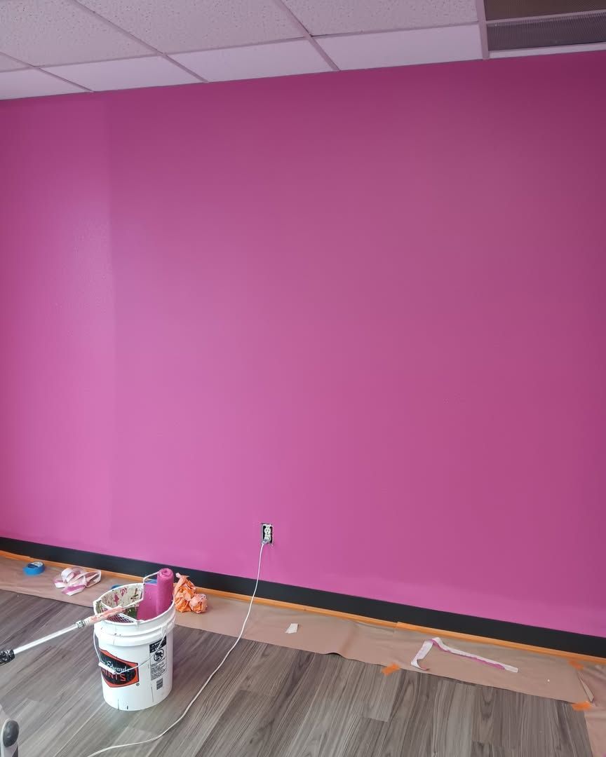 Bright pink wall in a room under renovation, with paint bucket, supplies, and exposed baseboard along the floor.