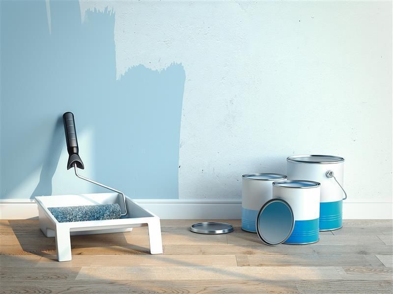 Paint roller, tray, and blue paint cans beside a half-painted light blue wall on a wooden floor.