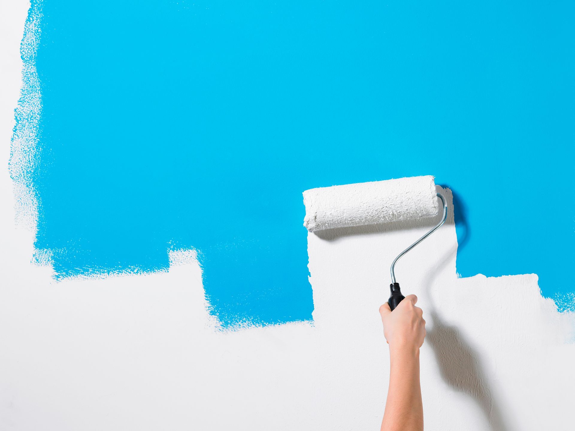 Hand using a paint roller to apply bright blue paint over a white wall