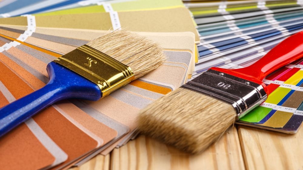 Two paintbrushes on paint color swatches, showing beige bristles and blue, gold, and red handles.