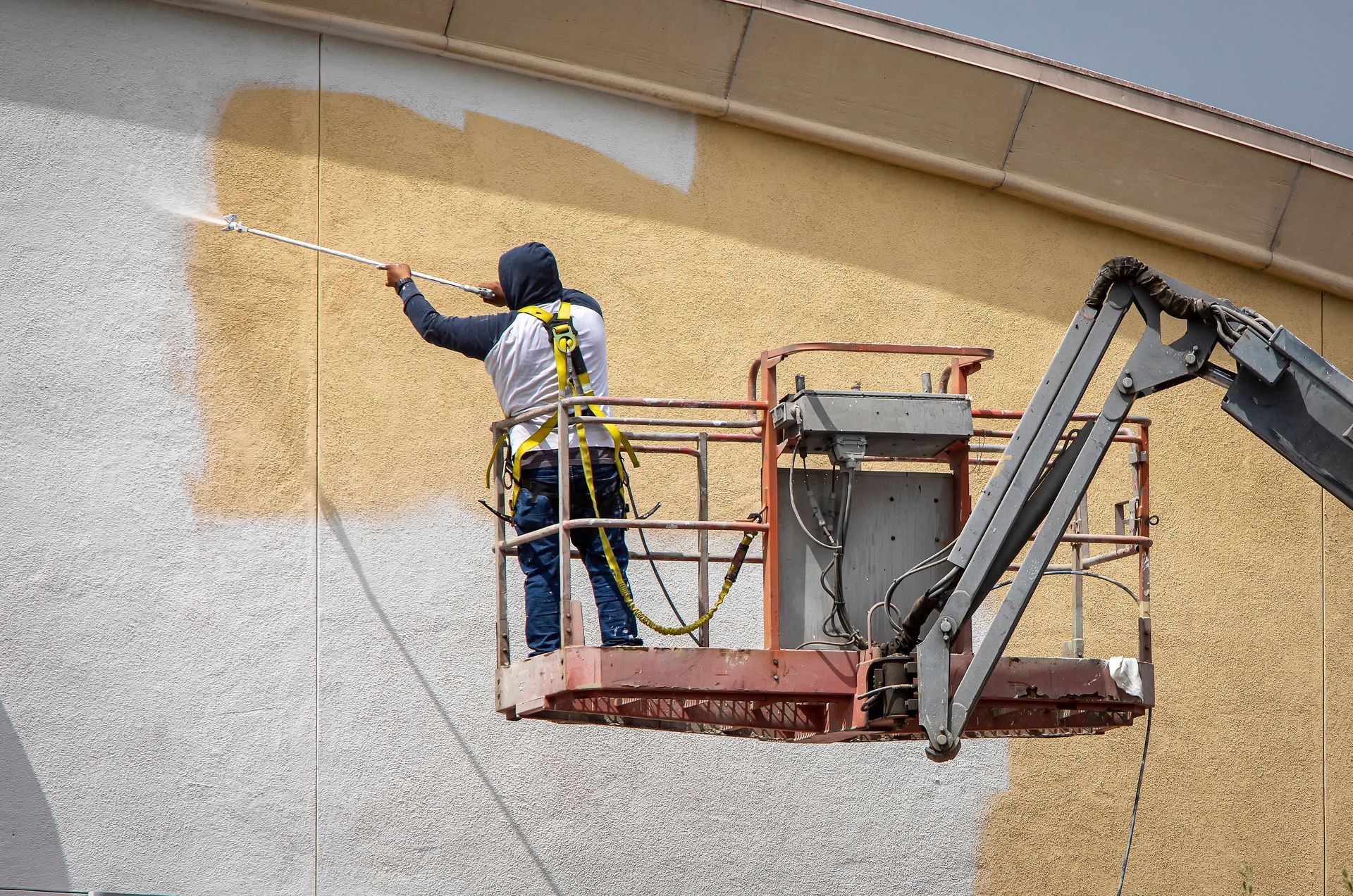 Worker on a lift spraying tan paint onto a building wall.