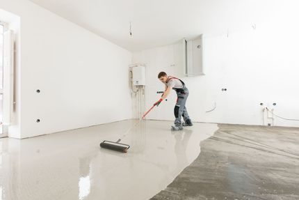 Worker smoothing wet concrete floor with a long-handled trowel in a bright, empty room